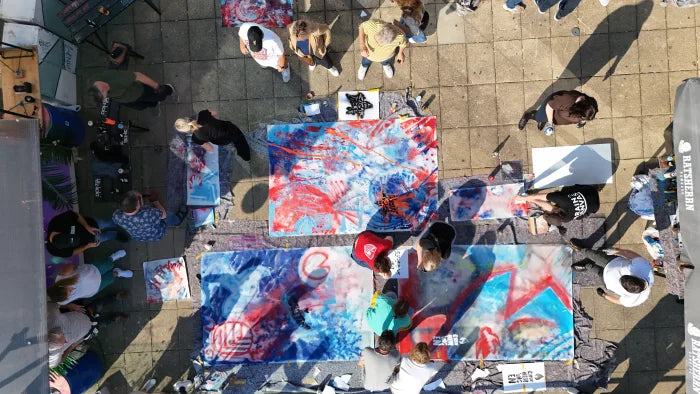 Luftaufnahme von oben: mehrere große Leinwände mit rot-weiß-blauen Action-Painting-Motiven liegen auf dem Boden, Teilnehmende arbeiten und stehen ringsum, Ratsherrn-Sonnenschirm am Rand