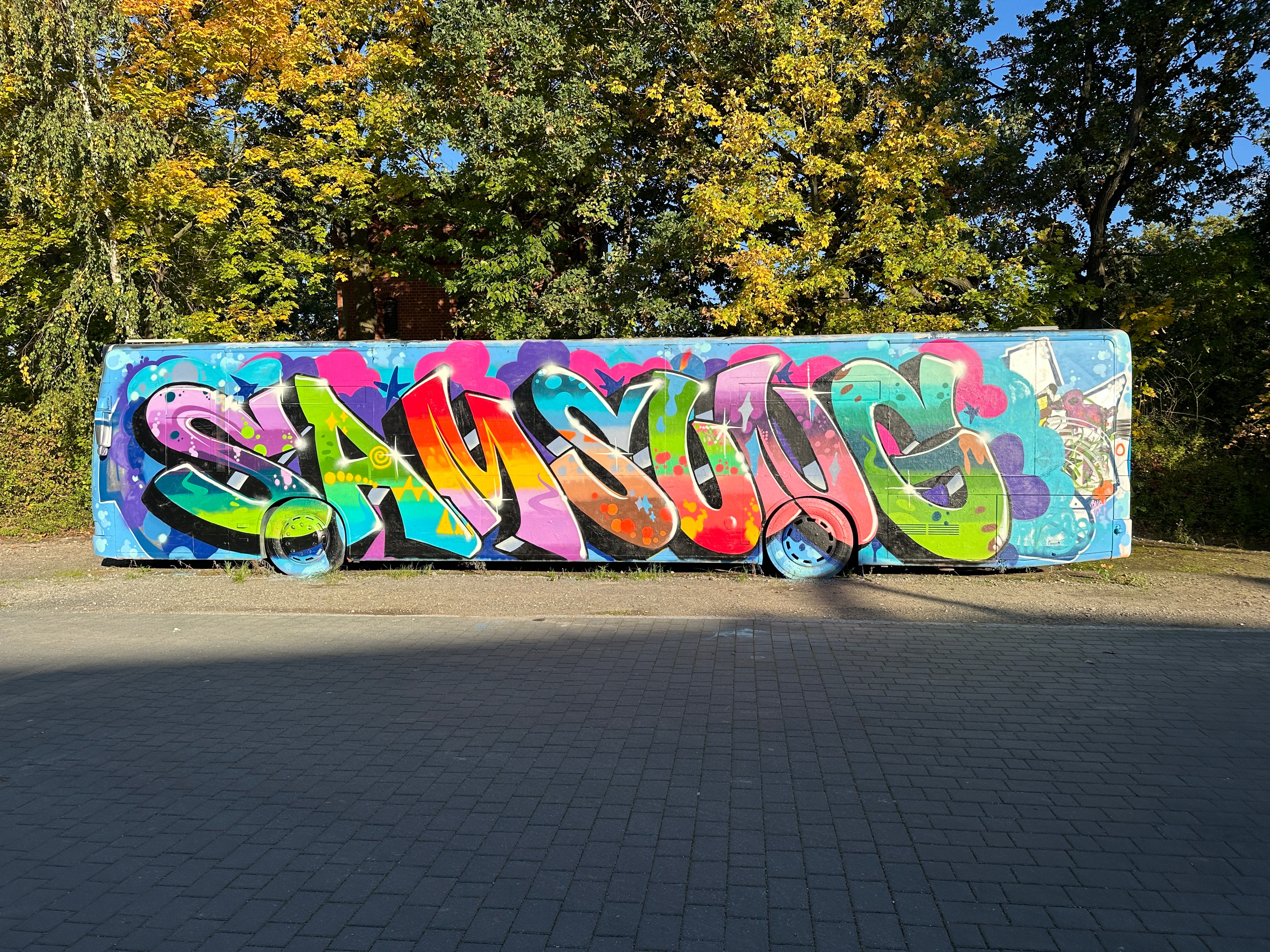 Kompletter Bus mit großem SAMSUNG-Schriftzug als Mural in Regenbogenfarben, umgeben von herbstlichen Bäumen im Wellenwerk