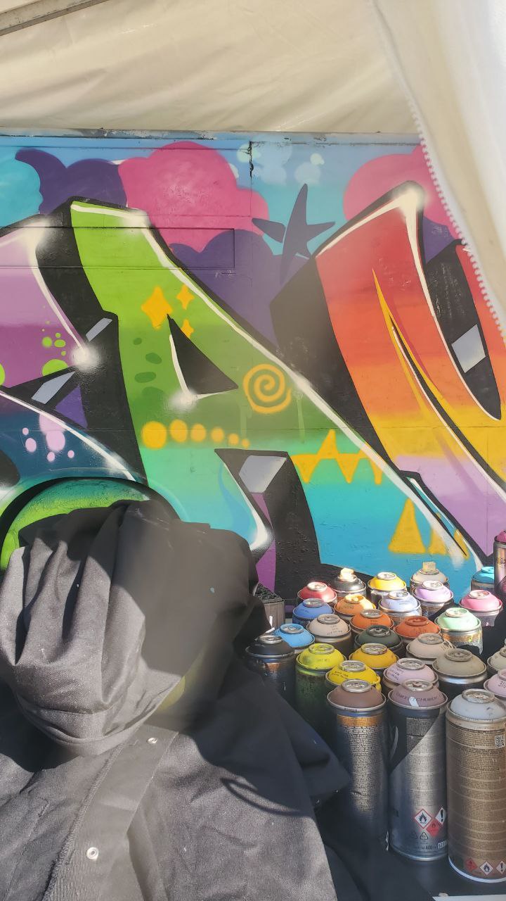 Nahaufnahme des Murals unter dem Zelt mit aufgereihten Spraydosen im Vordergrund, sehr detailliert gearbeitete Farbverläufe