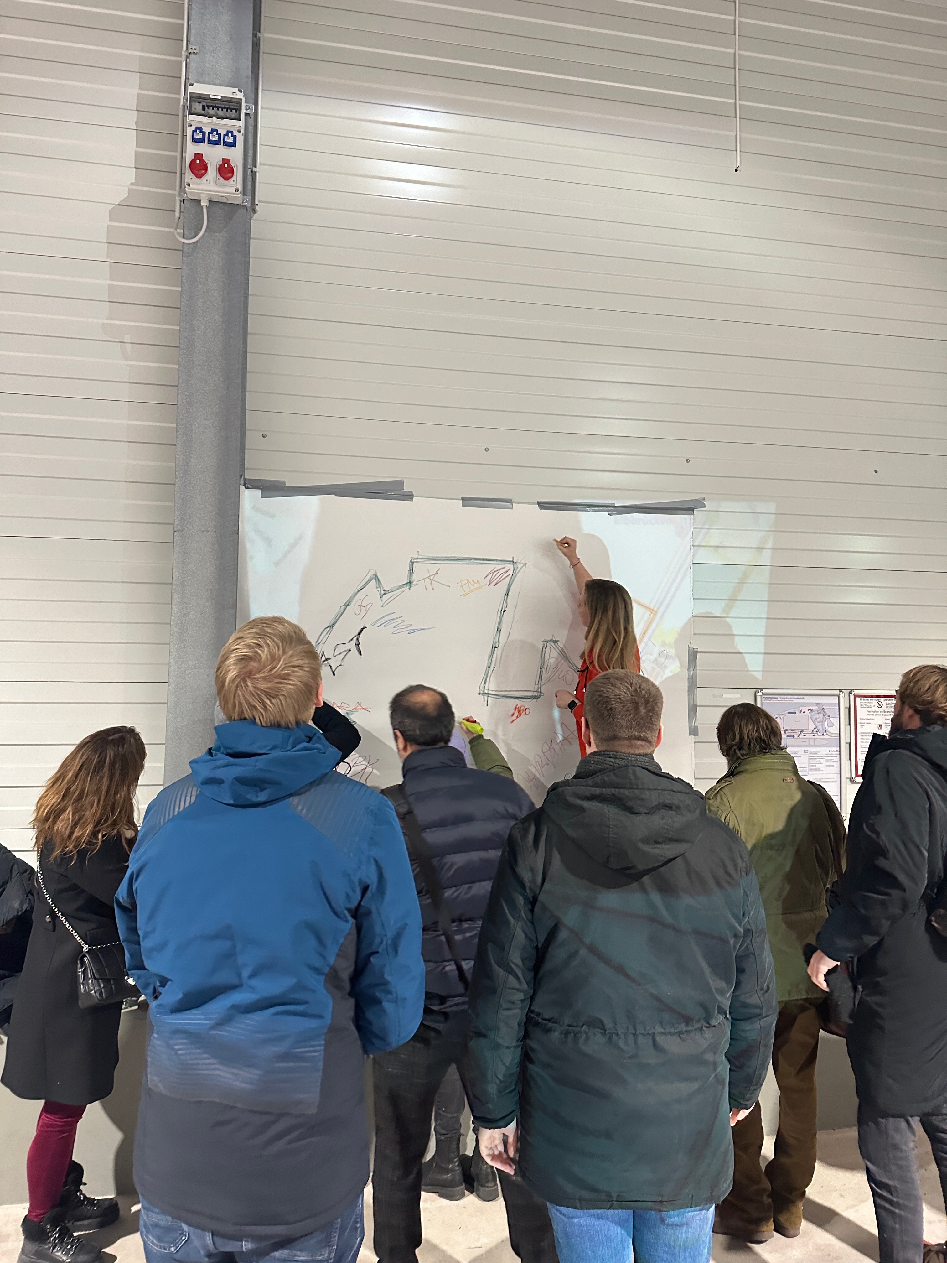 Gruppe steht vor einer an die Wand geklebten Papierbahn und skizziert gemeinsam einen ersten Entwurf