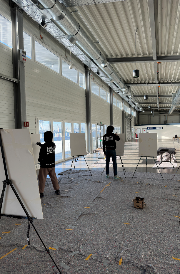Zwei Personen in Brain2Canvas-Hoodies stehen an leeren Staffeleien in einer großen Halle mit Fensterfront zum Hafen