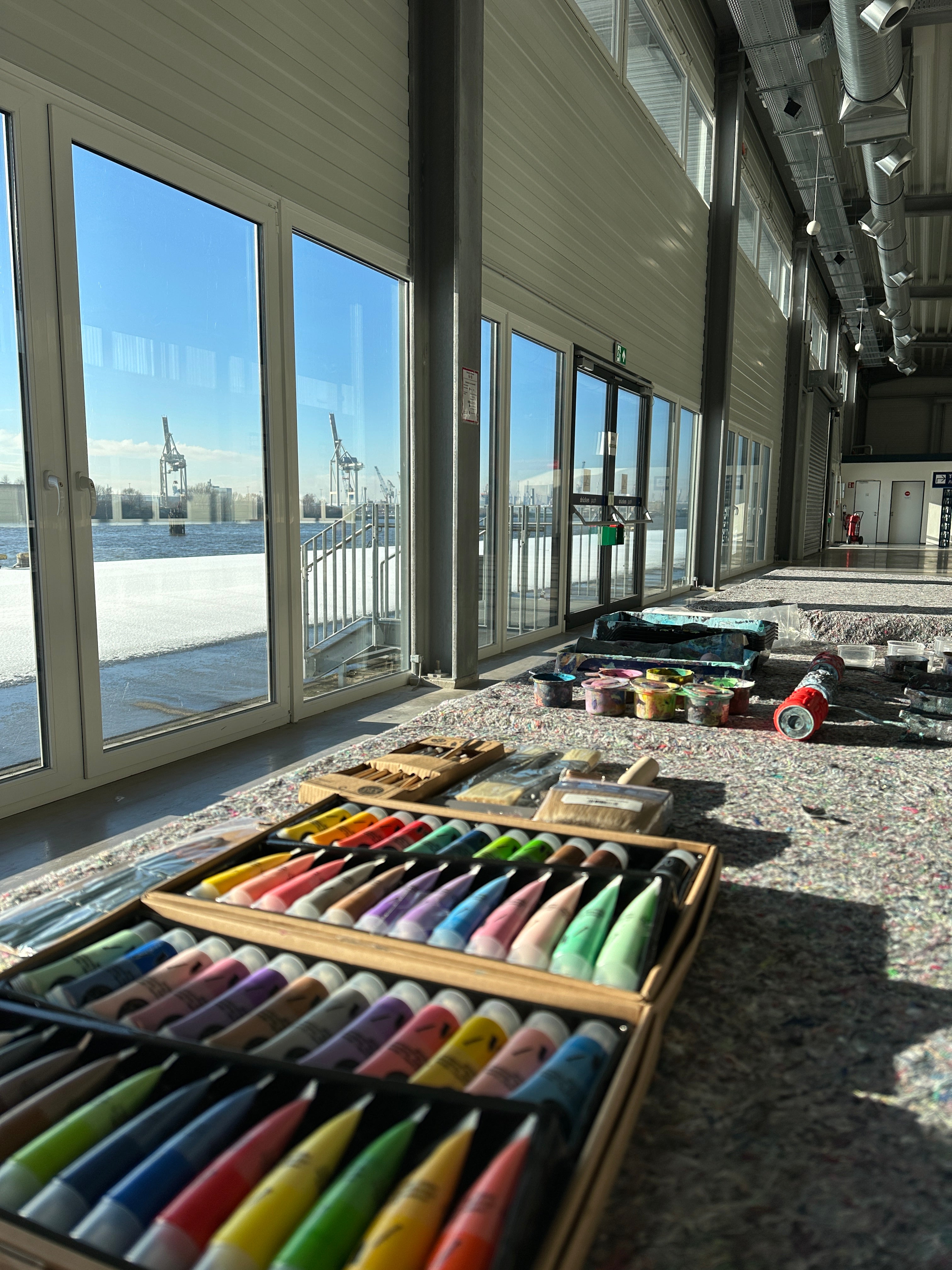 Malutensilien und Farbtuben liegen auf dem Boden einer Terminal-Halle in der HafenCity, durch die Fensterfront Blick auf die Elbe mit Hafenkränen