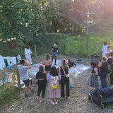 Abendaufnahme des Lingoda-Sommerfests: Gaeste stehen um eine Leinwand am Boden, links haengen bemalte Leinwaende